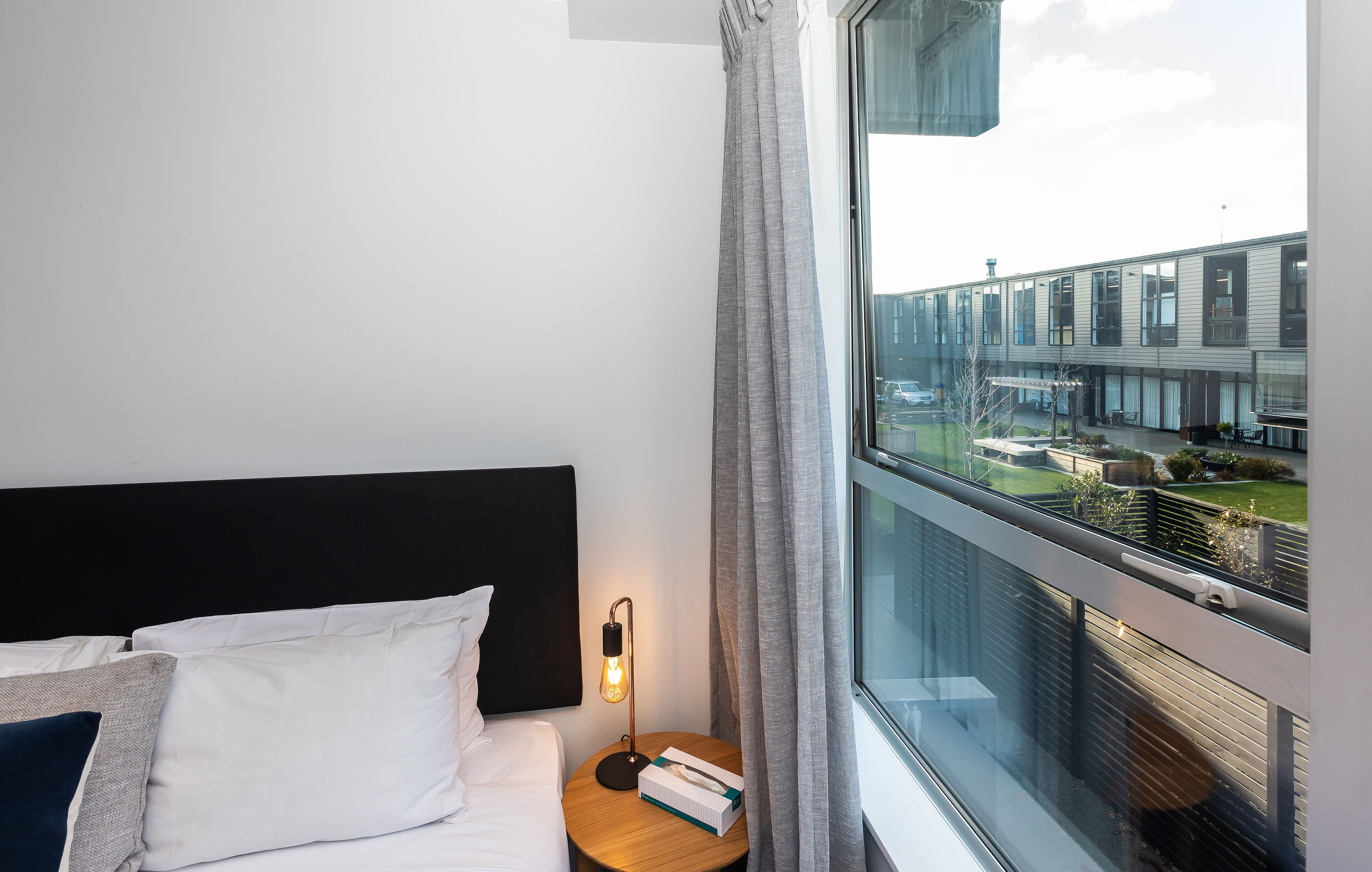 A hotel room with a bed, a bedside table with a lamp, and a window overlooking a building.