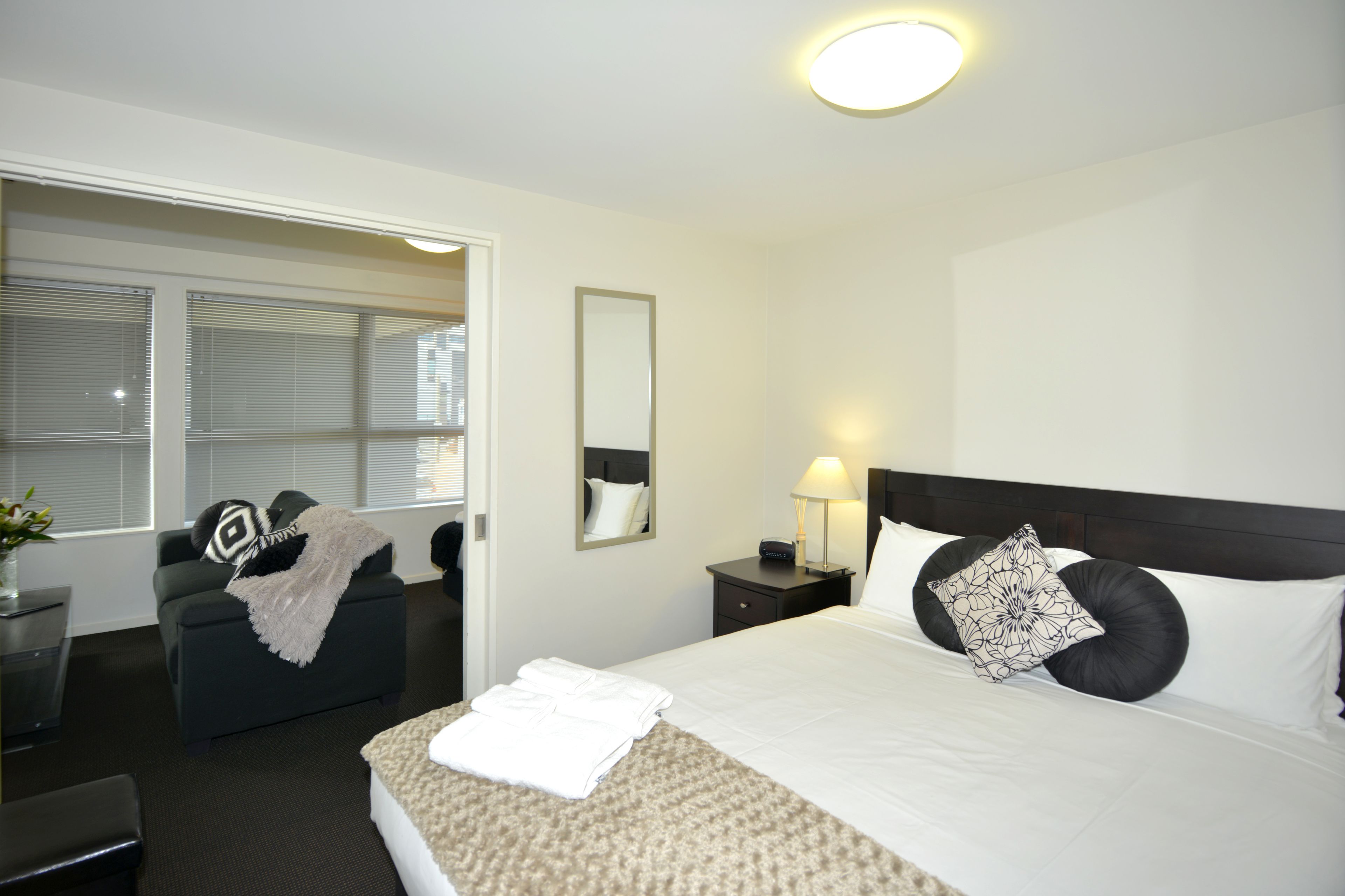 A hotel room with a bed, a desk, a chair, a nightstand, a lamp, a mirror, and a window with blinds.