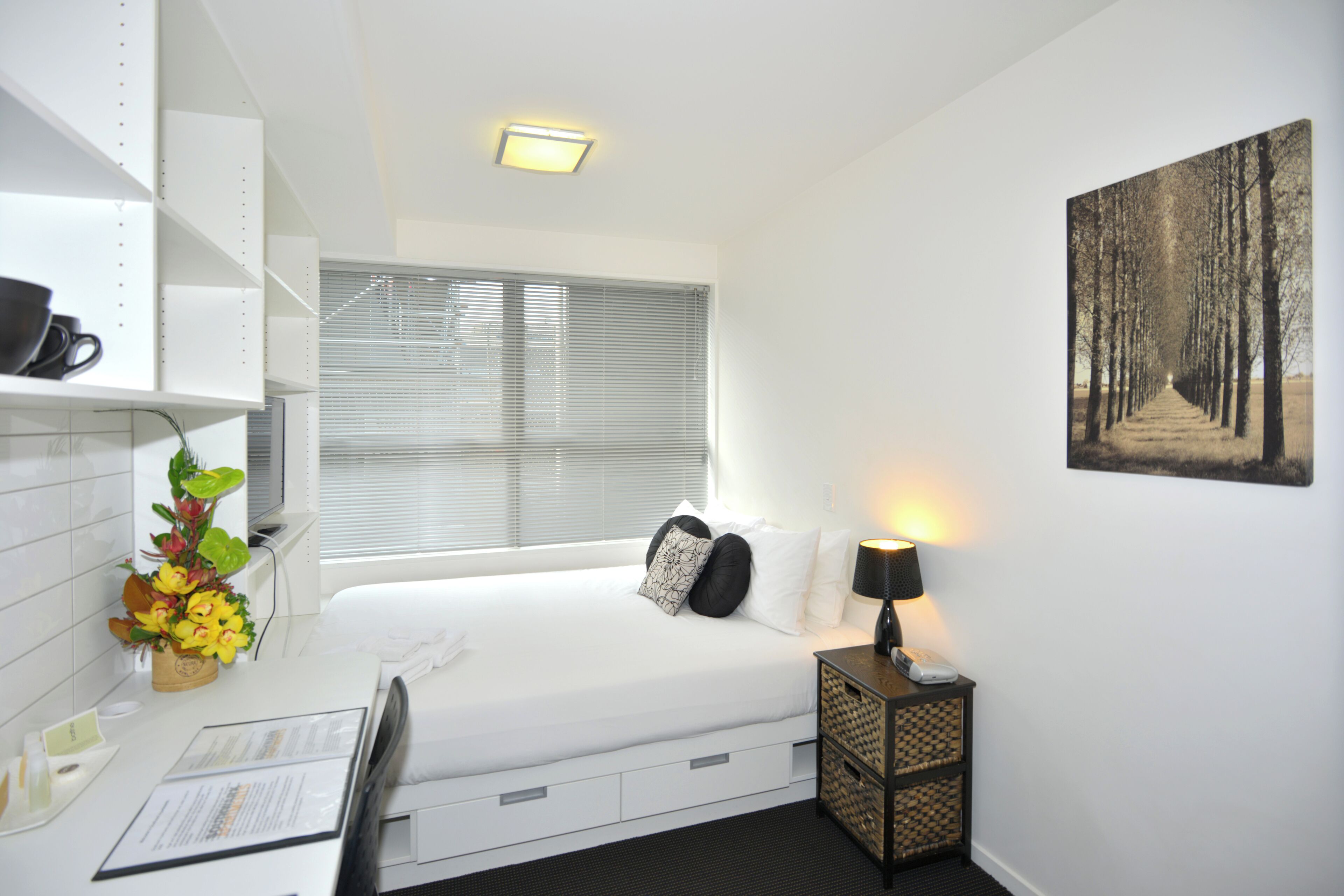 A modern hotel room with a neatly made bed, a bedside table with a lamp, a window with blinds, a wall-mounted shelf with a coffee cup, and a framed picture on the wall.