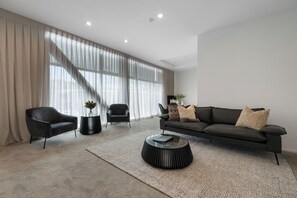 Een moderne zithoek met een bank, twee fauteuils, een salontafel en een groot raam met lichte gordijnen.