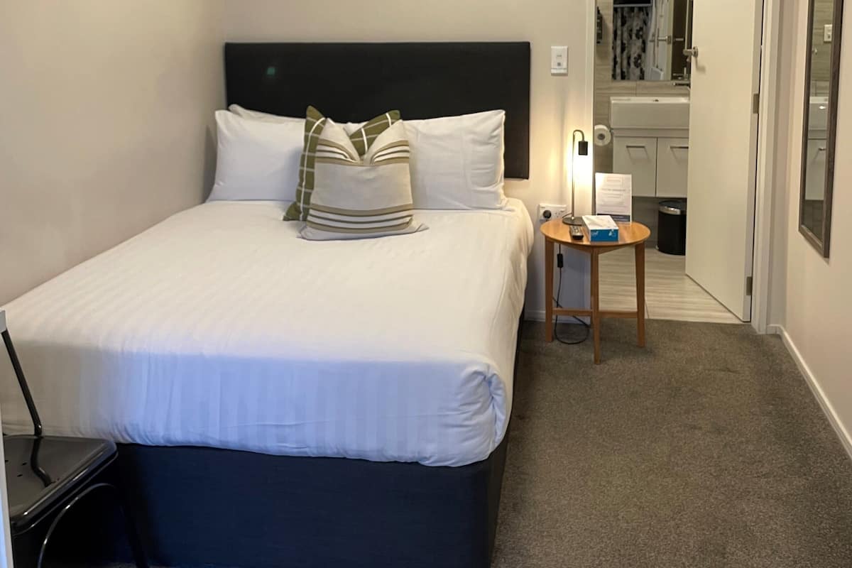 Un lit bien fait, avec du linge de lit blanc et une tête de lit sombre, une table de chevet avec une lampe et un petit miroir accroché au mur.