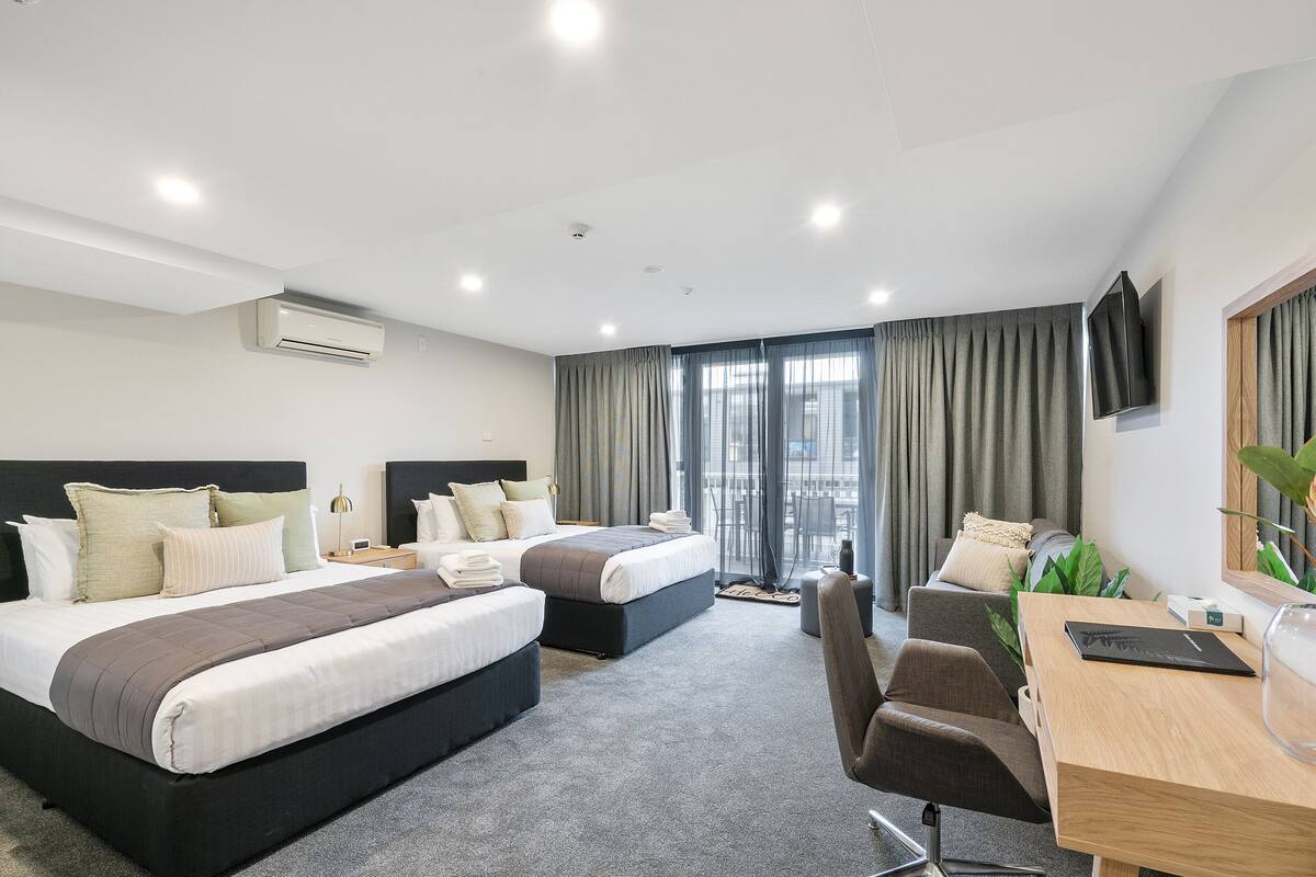 Une chambre d’hôtel avec deux lits, un bureau, une chaise, une télévision et une grande fenêtre avec des rideaux.
