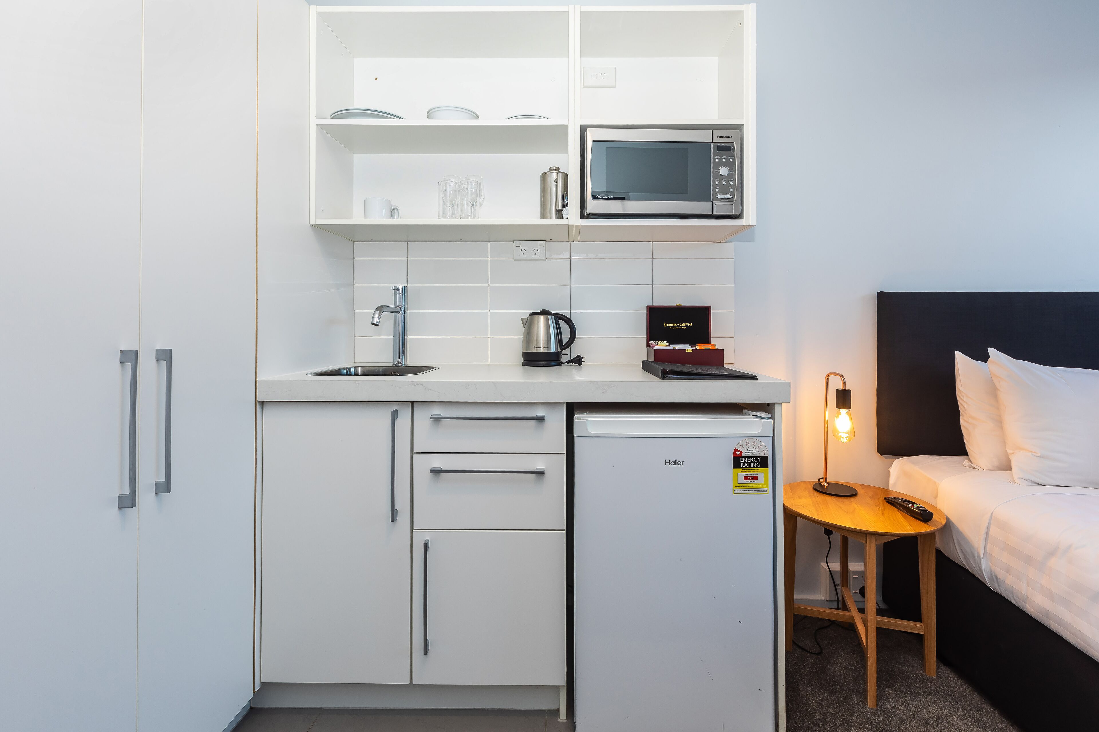 Una piccola cucina compatta con forno a microonde, bollitore ed un piccolo frigorifero. Un letto con biancheria bianca e un comodino con una lampada.