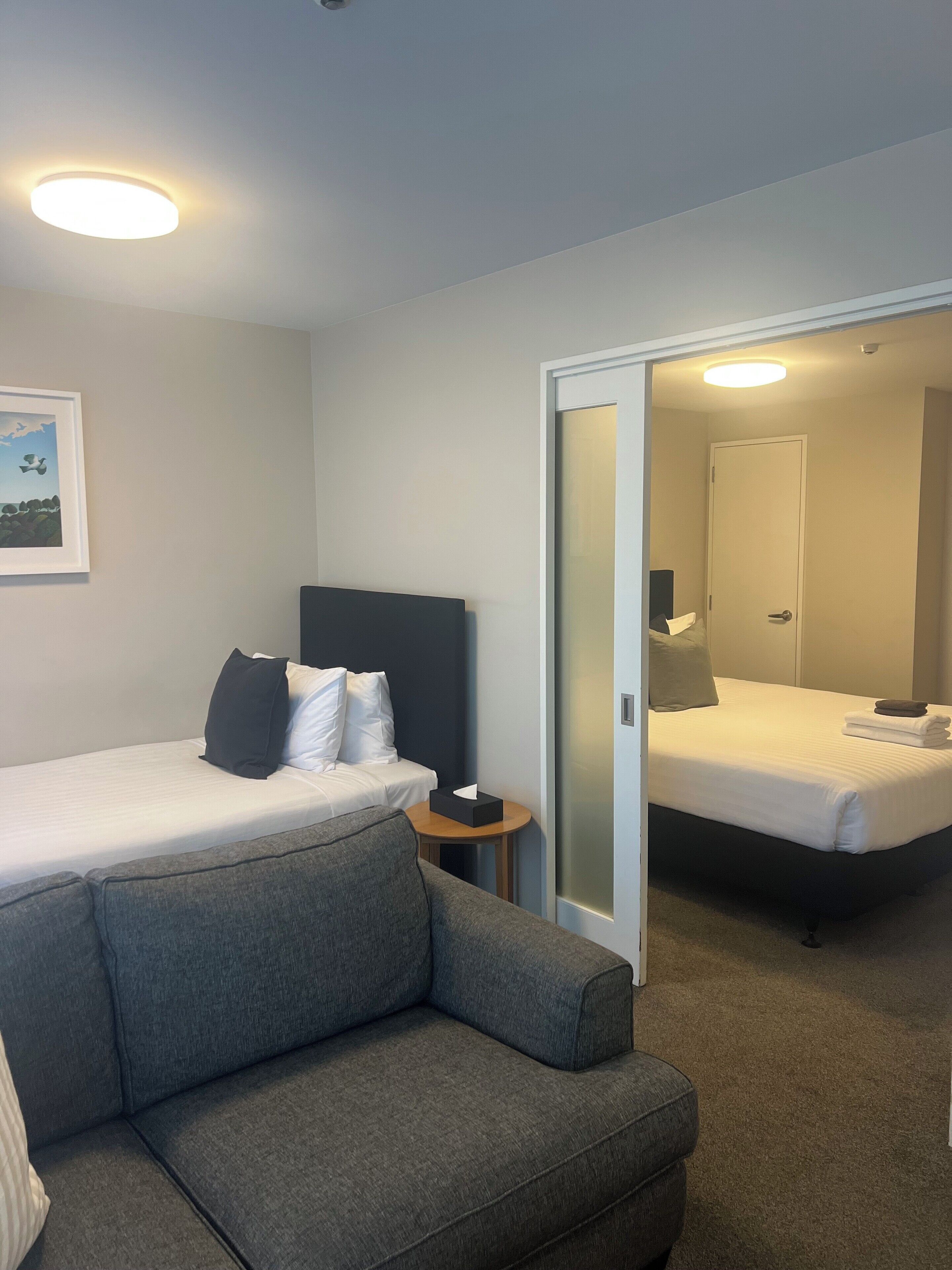 Habitació d'hotel amb un llit, un sofà, una tauleta de nit i una porta que dona a una altra habitació.
