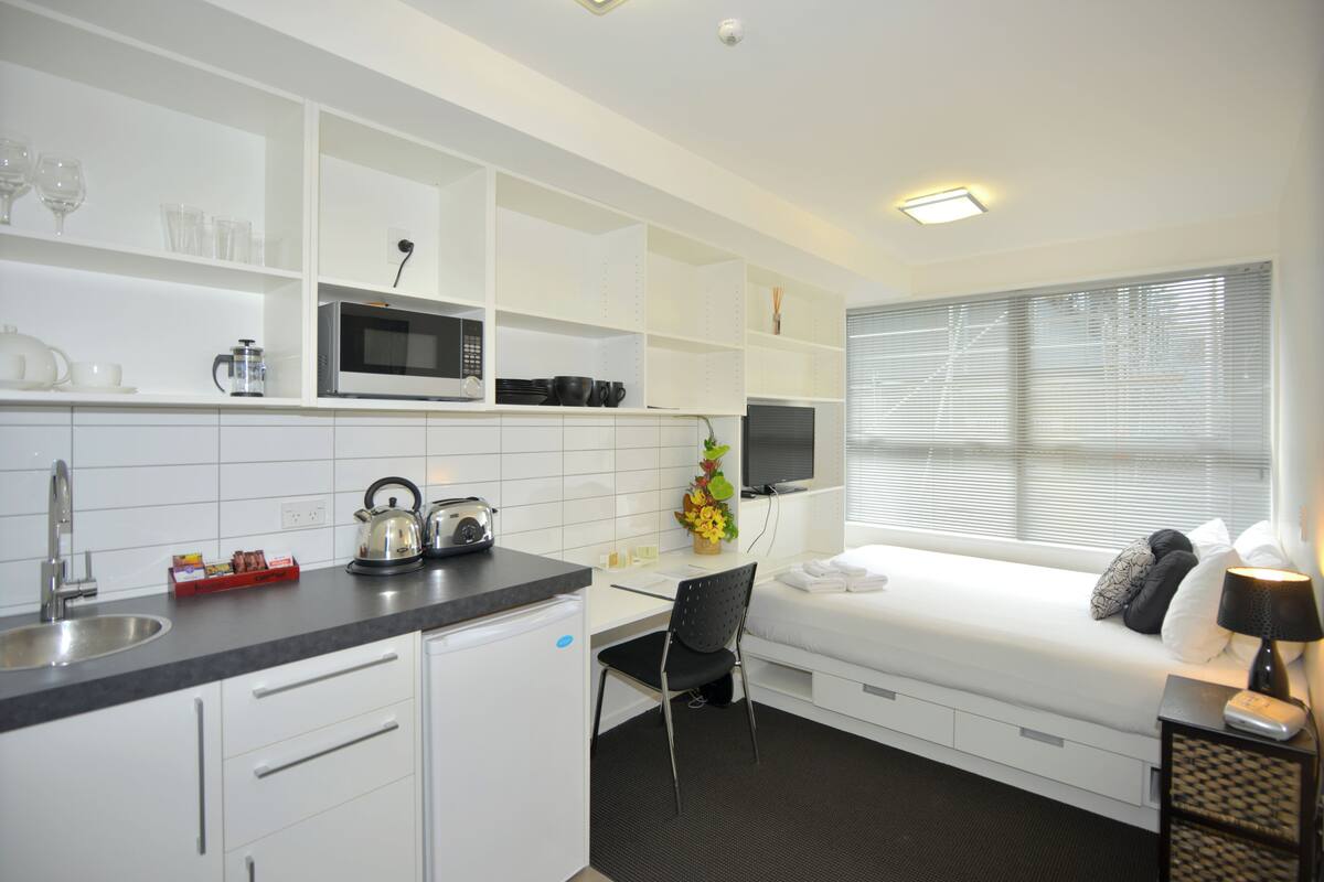 Une chambre d’hôtel compacte comprenant un coin cuisine, un lit avec des oreillers, un bureau avec une chaise et une fenêtre avec des stores.