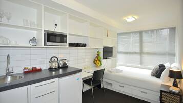 A compact hotel room with a kitchenette, a bed with pillows, a desk with a chair, and a window with blinds.