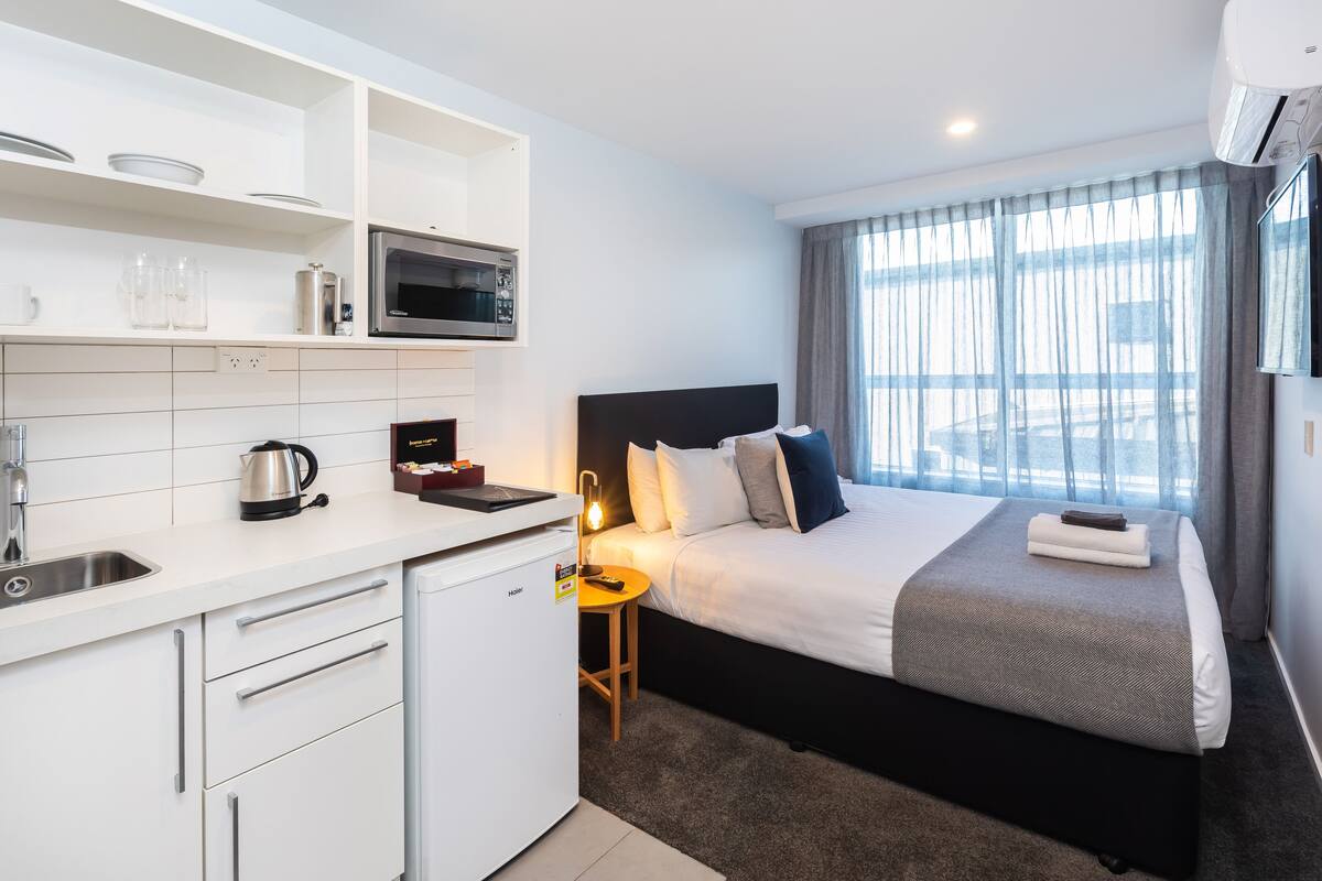 Une chambre d’hôtel moderne dotée d’une kitchenette, d’un lit avec une tête de lit, d’une table de chevet avec une lampe et d’une grande fenêtre avec des rideaux.