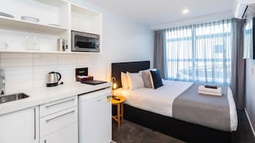 Una habitación de hotel moderna con kitchenette, una cama con cabecera, una mesita de noche con lámpara y un ventanal con cortinas.