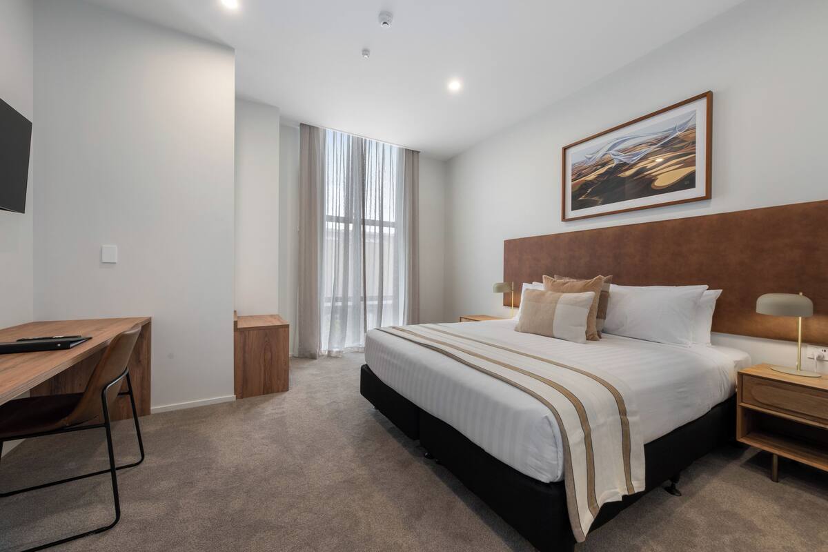 Une chambre d’hôtel moderne avec un grand lit, un bureau et une fenêtre ornée de rideaux légers.
