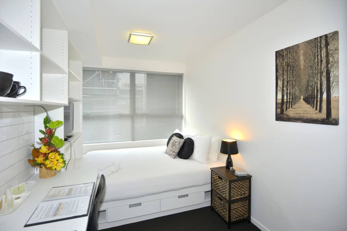 Une chambre d’hôtel compacte comprenant un lit, une lampe de chevet, un petit bureau avec une chaise, une fenêtre avec des stores, une étagère fixée au mur avec une tasse à café, et un tableau encadré accroché au mur.