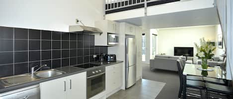 A modern kitchen with a dishwasher, oven, and double sink. There is a dining area with a table and chairs. A balcony is visible above the kitchen.