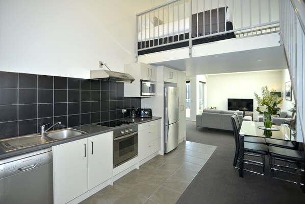 A modern kitchen with a dishwasher, oven, and double sink. There is a dining area with a table and chairs. A balcony is visible above the kitchen.