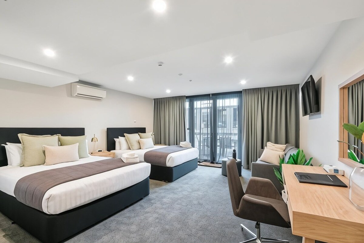 A hotel room with two beds, a desk, a chair, a TV, and a large window with curtains.
