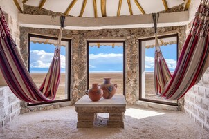 Una habitación con pared de piedra, un banco, dos macetas de barro y vistas a una playa a través de dos ventanas.