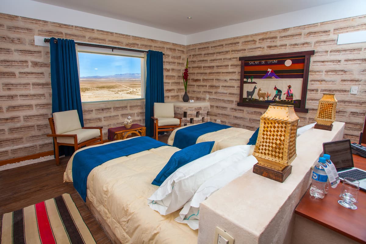 Quarto de hotel com duas camas, uma mesa com um laptop, uma cadeira, uma mesinha de cabeceira, um abajur decorativo e um quadro na parede.