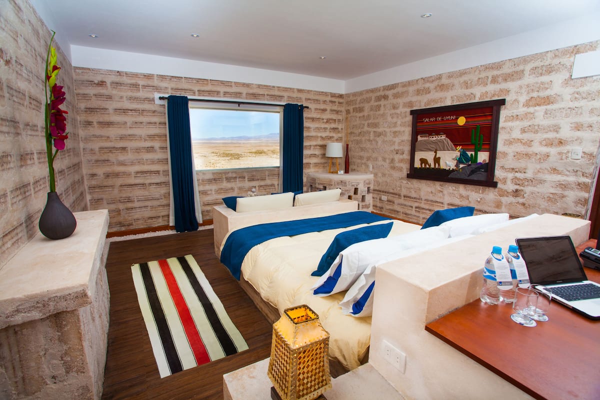Quarto de hotel com cama, escrivaninha com laptop, vaso com flores, janela com vista para o deserto e parede de tijolos.