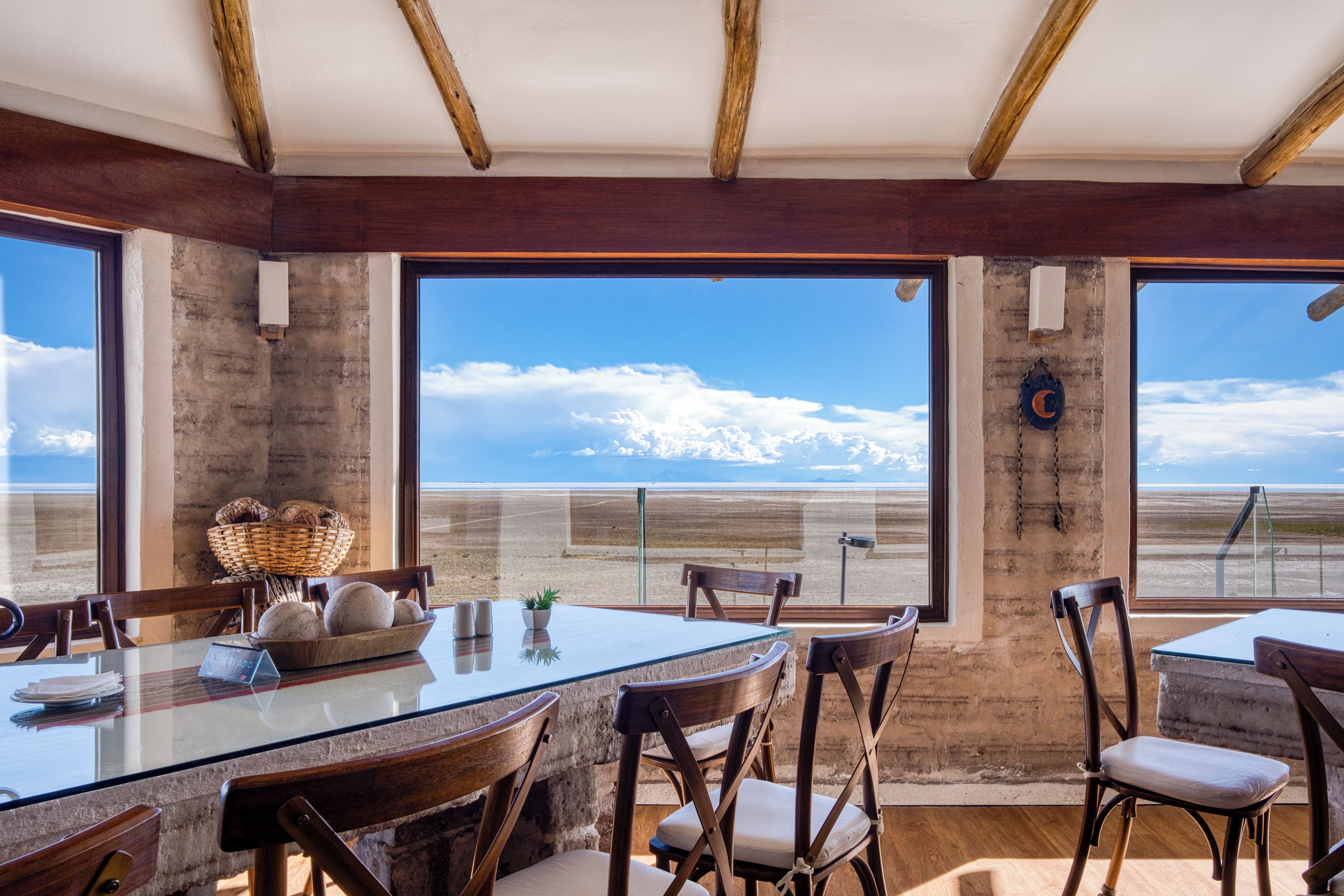 Un espace repas avec une table en verre et des chaises en bois, de grandes fenêtres offrant une vue sur la plage et les montagnes, et un intérieur rustique aux murs en béton apparent.