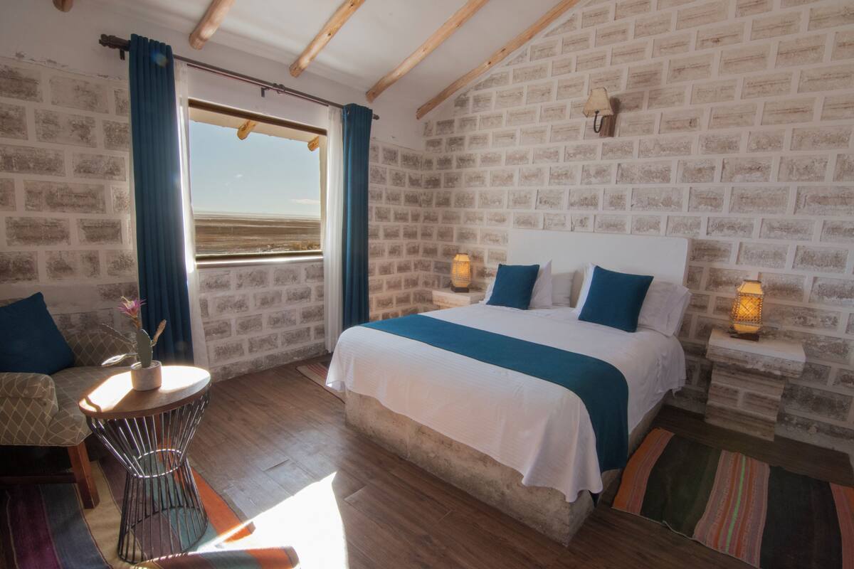 Quarto com uma cama grande, uma mesinha com uma planta, uma cadeira e vista para a praia pela janela.