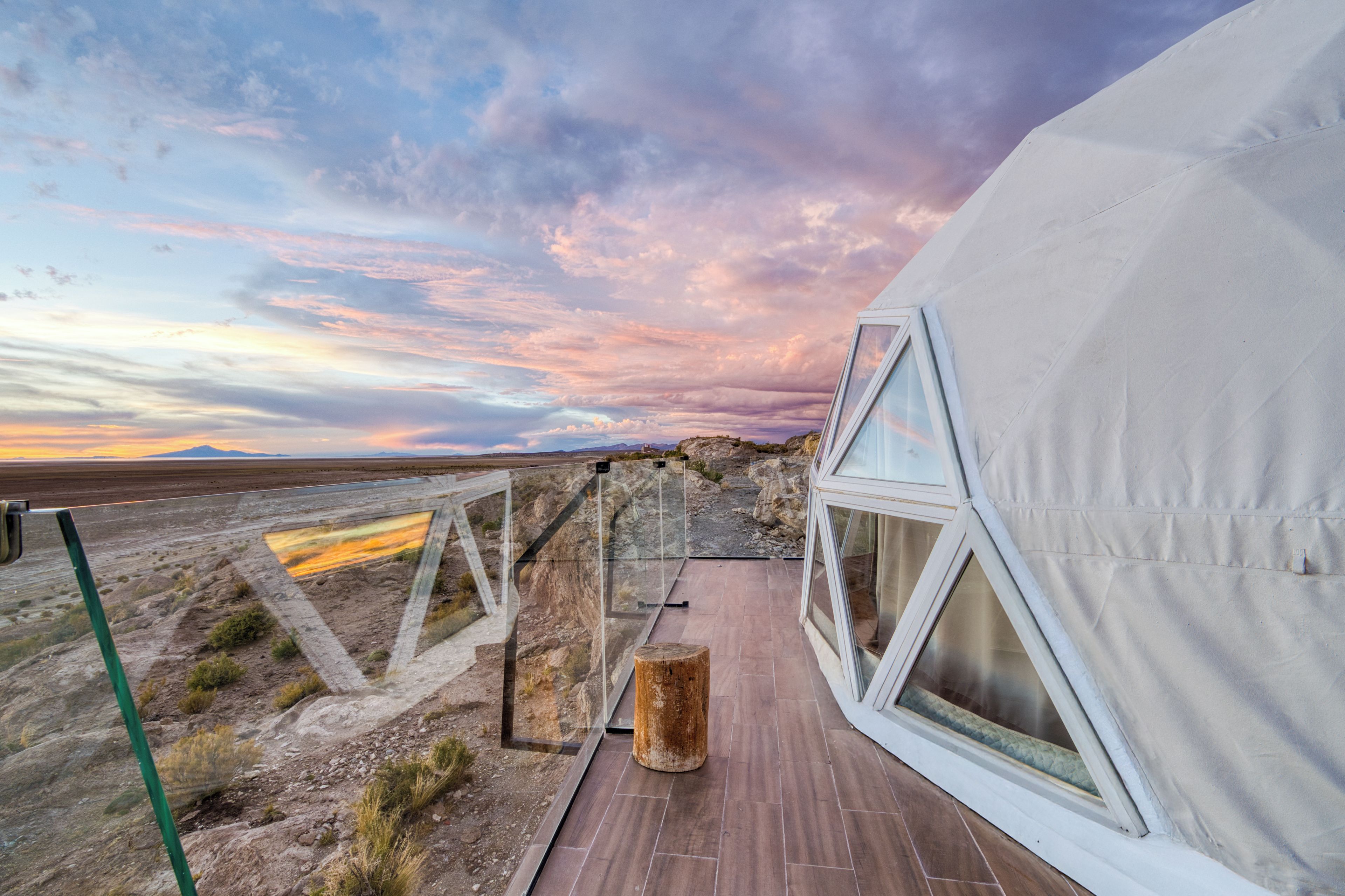 A modern tent-like structure with a wooden deck and large windows, set against a backdrop of a desert landscape and a colorful sky.