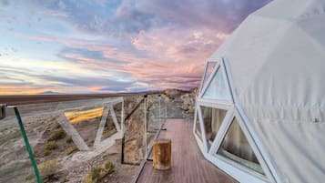 A modern tent-like structure with a wooden deck and large windows, set against a backdrop of a desert landscape and a colorful sky.