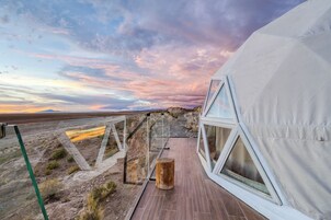 Una estructura moderna tipo tienda de campaña con terraza de madera y ventanas amplias, ubicada frente a un paisaje desértico y un cielo colorido.