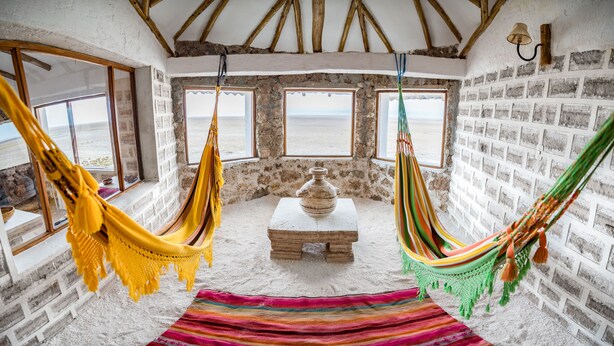 A room with a rustic stone wall, a hammock, a colorful striped rug, and a view of the sea through large windows.