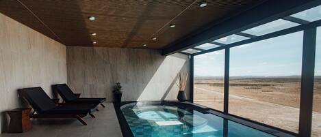 A modern indoor pool area with two lounge chairs and a large window overlooking a desert landscape.
