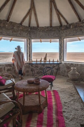 Una habitación con techo alto, vigas de madera rústicas, un ventanal con vistas a un paisaje desértico, una mesa con sillas y un tapete de fibras naturales.