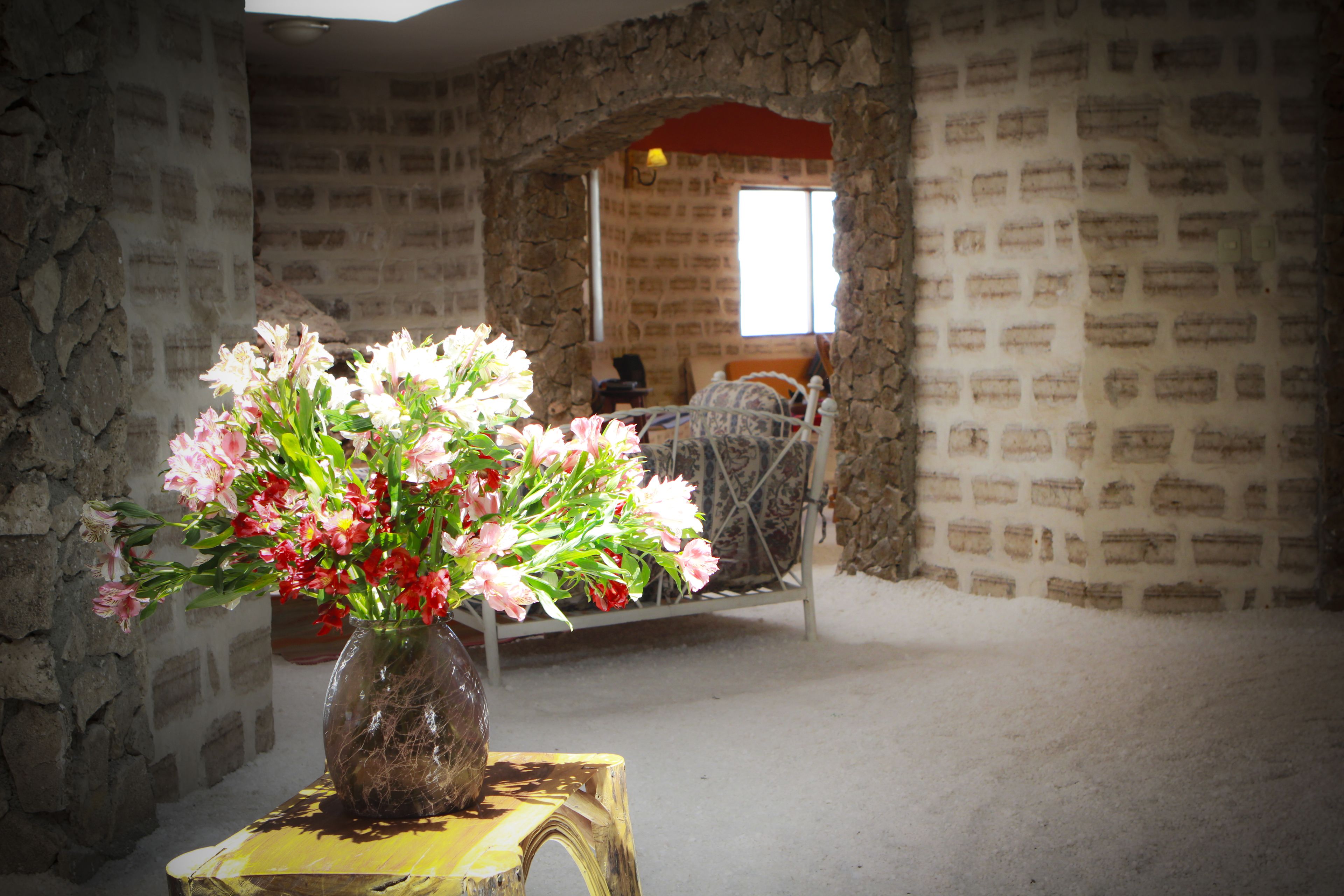 Una habitación con pared de piedra, cama con estructura de metal y un florero con flores sobre un taburete de madera.