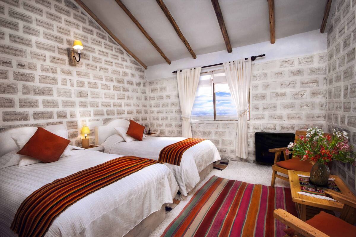 Quarto com duas camas, uma mesa de madeira com um vaso e uma janela com cortinas.