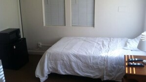 Un dormitorio con cama, dos ventanas con persianas, una lámpara, un escritorio con computadora y un televisor.