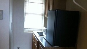 Una pequeña cocina con armarios de madera, un refrigerador negro, una ventana con persianas y un cubo de basura.