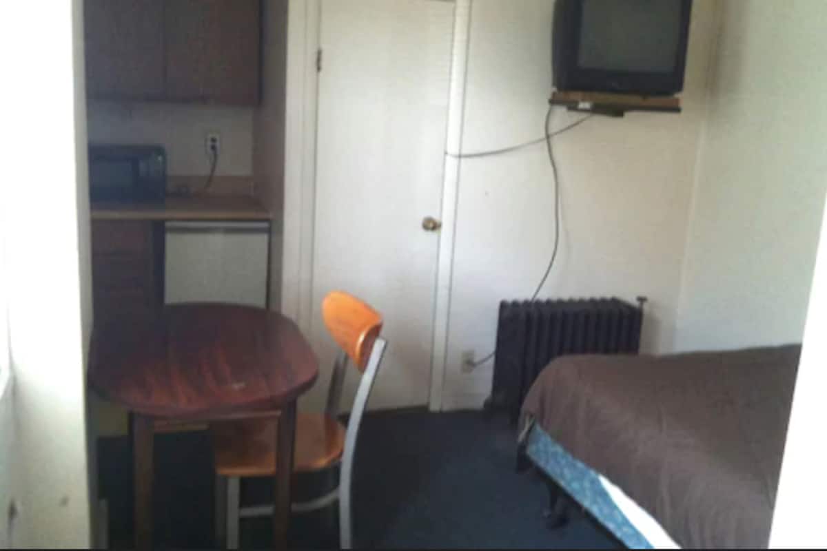 A small room with a bed, a chair, a table, a microwave, and a television mounted on the wall.