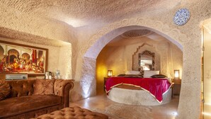 A luxurious room with a plush brown sofa, a tufted ottoman, a large bed with a red bedspread, and a painting on the wall. - Ottoman Cave Suites (Nevsehir)