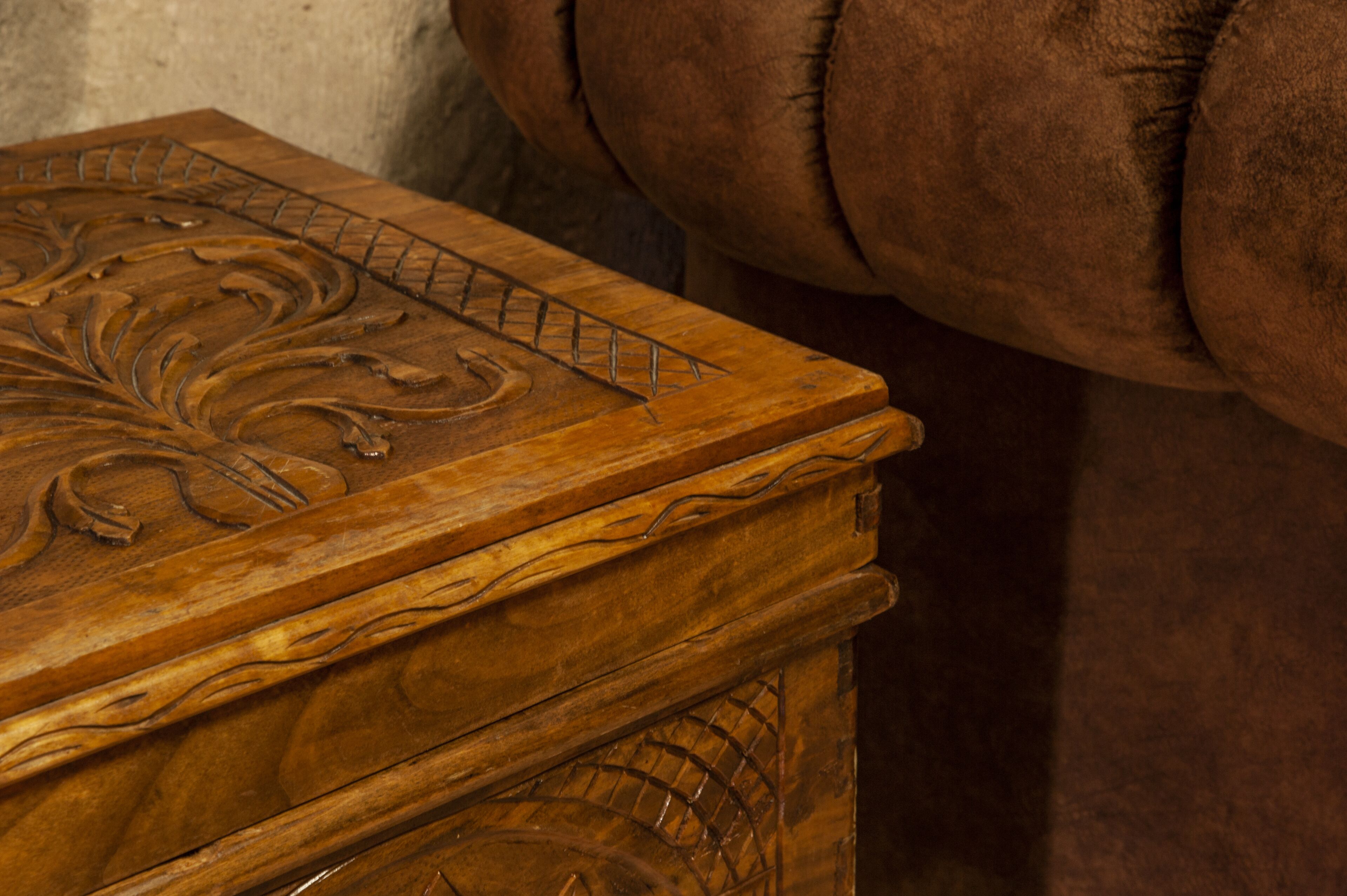 A wooden chest with intricate carvings, positioned next to a brown upholstered chair.