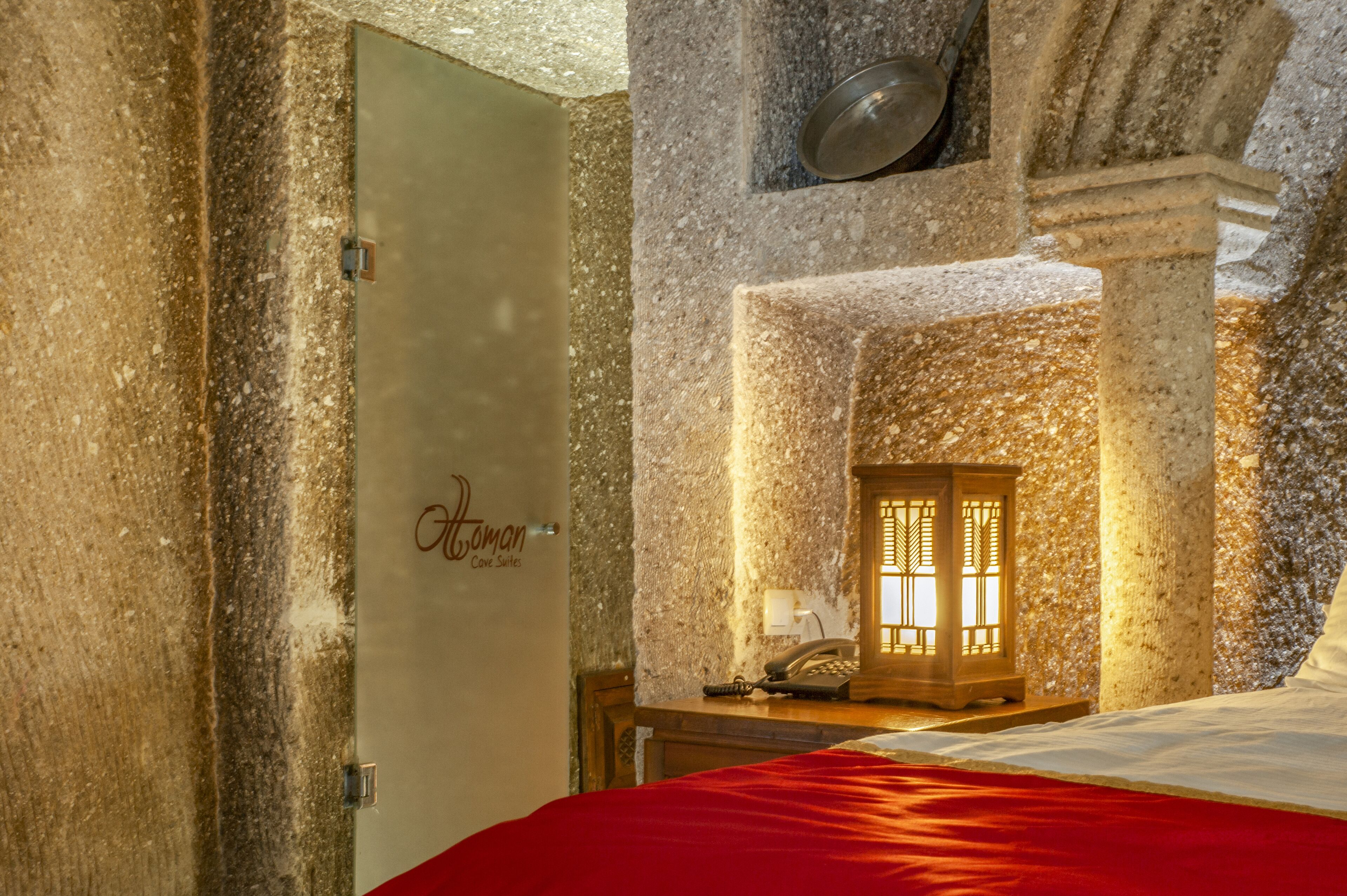 A hotel room with a stone wall, a bed with a red bedspread, a wooden nightstand with a lamp, and a door with a sign.