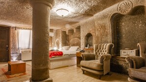 A room with a large stone column, a bed with a red blanket, two upholstered chairs, a wooden chest, and a patterned rug. - Ottoman Cave Suites (Nevsehir)