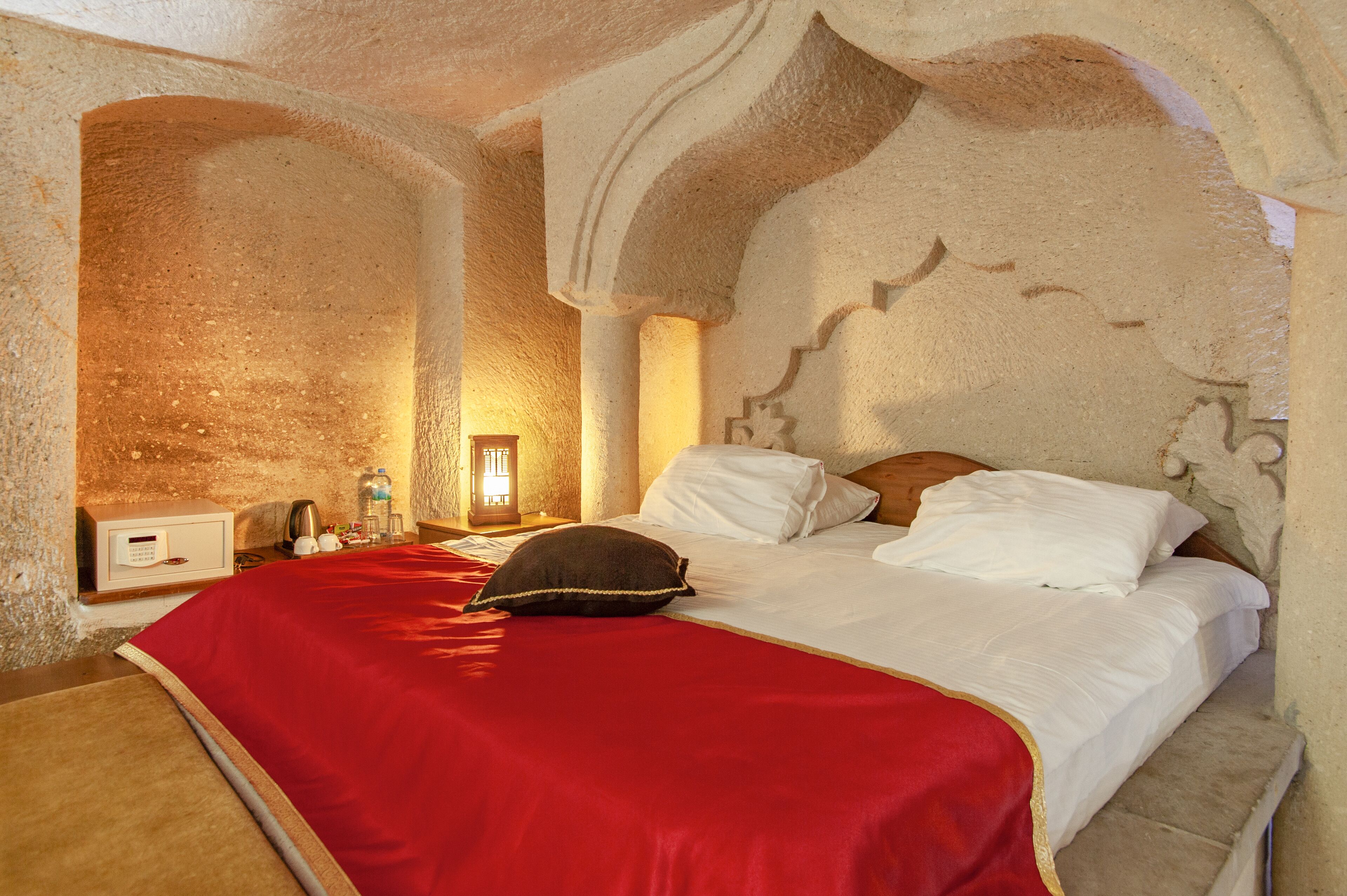A room with a large bed, a red blanket, and a wooden headboard. There is a nightstand with a lamp and a small table with a bottle and a cup. The walls have a unique stone texture.