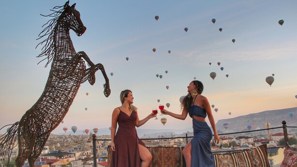 A terrace with a decorative horse sculpture, a table with food and drinks, and a view of a cityscape with hot air balloons in the sky. - Ottoman Cave Suites (Nevsehir)