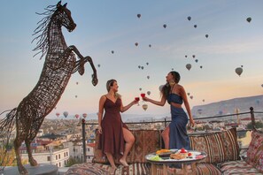 A terrace with a decorative horse sculpture, a table with food and drinks, and a view of a cityscape with hot air balloons in the sky. - Ottoman Cave Suites (Nevsehir)
