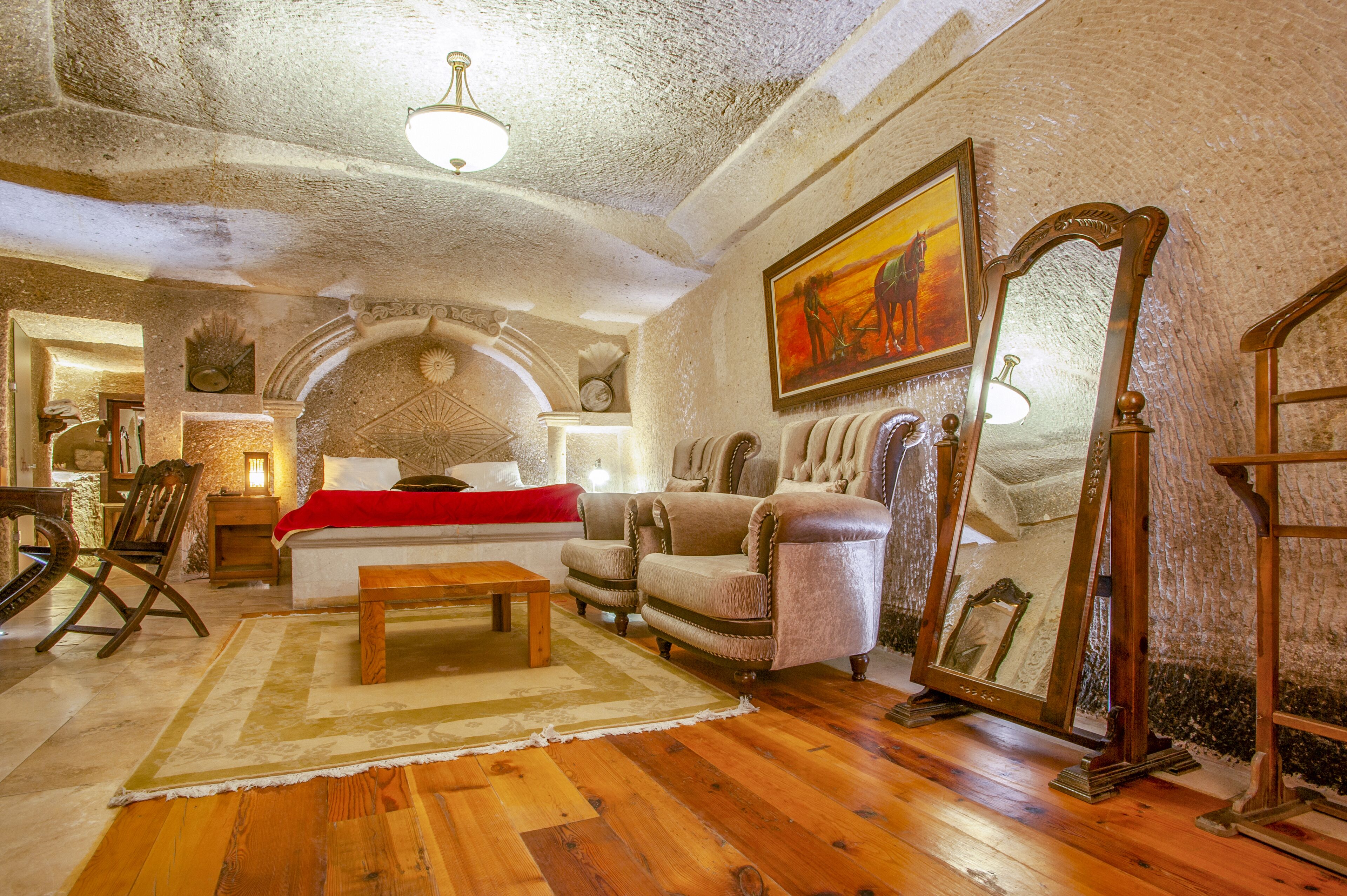 A cozy room with a wooden floor, a bed with a red blanket, two armchairs, a wooden coffee table, and a large mirror.