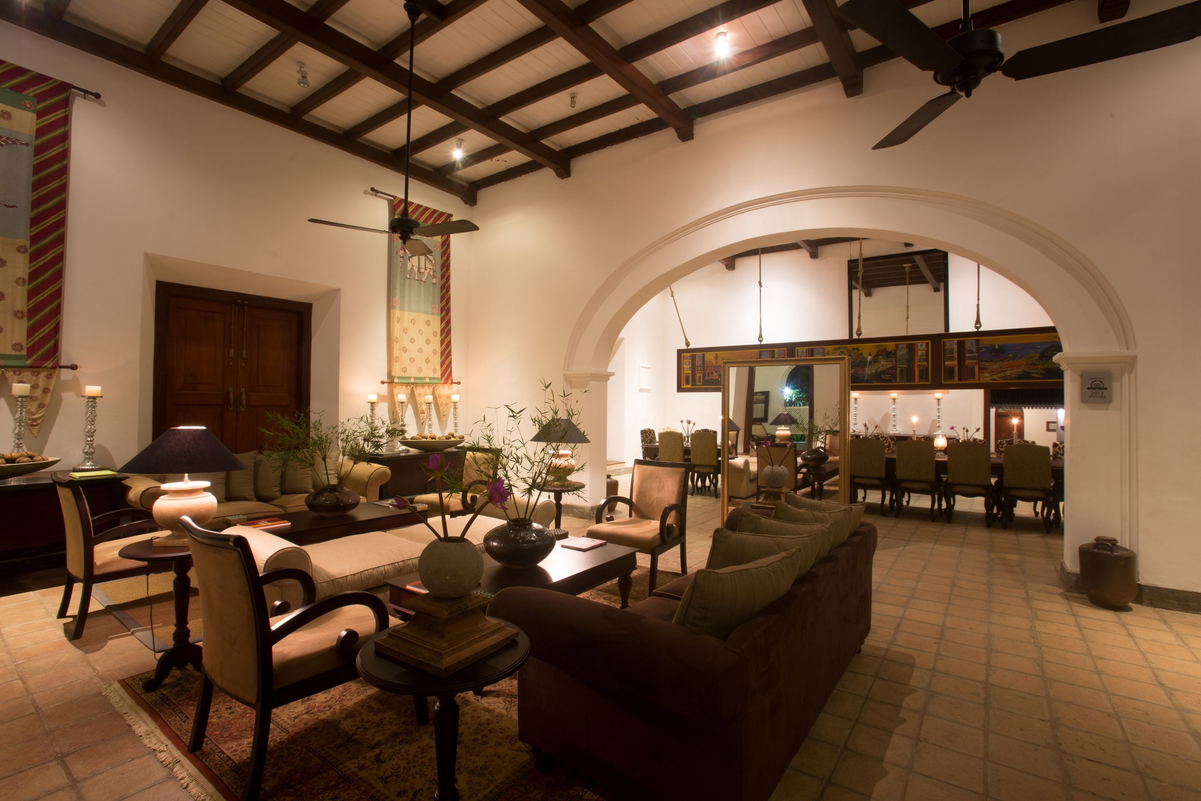 A spacious living room with a high ceiling, wooden beams, and a large arched doorway leading to another room. There are multiple seating areas with sofas and chairs, and a coffee table in the center.