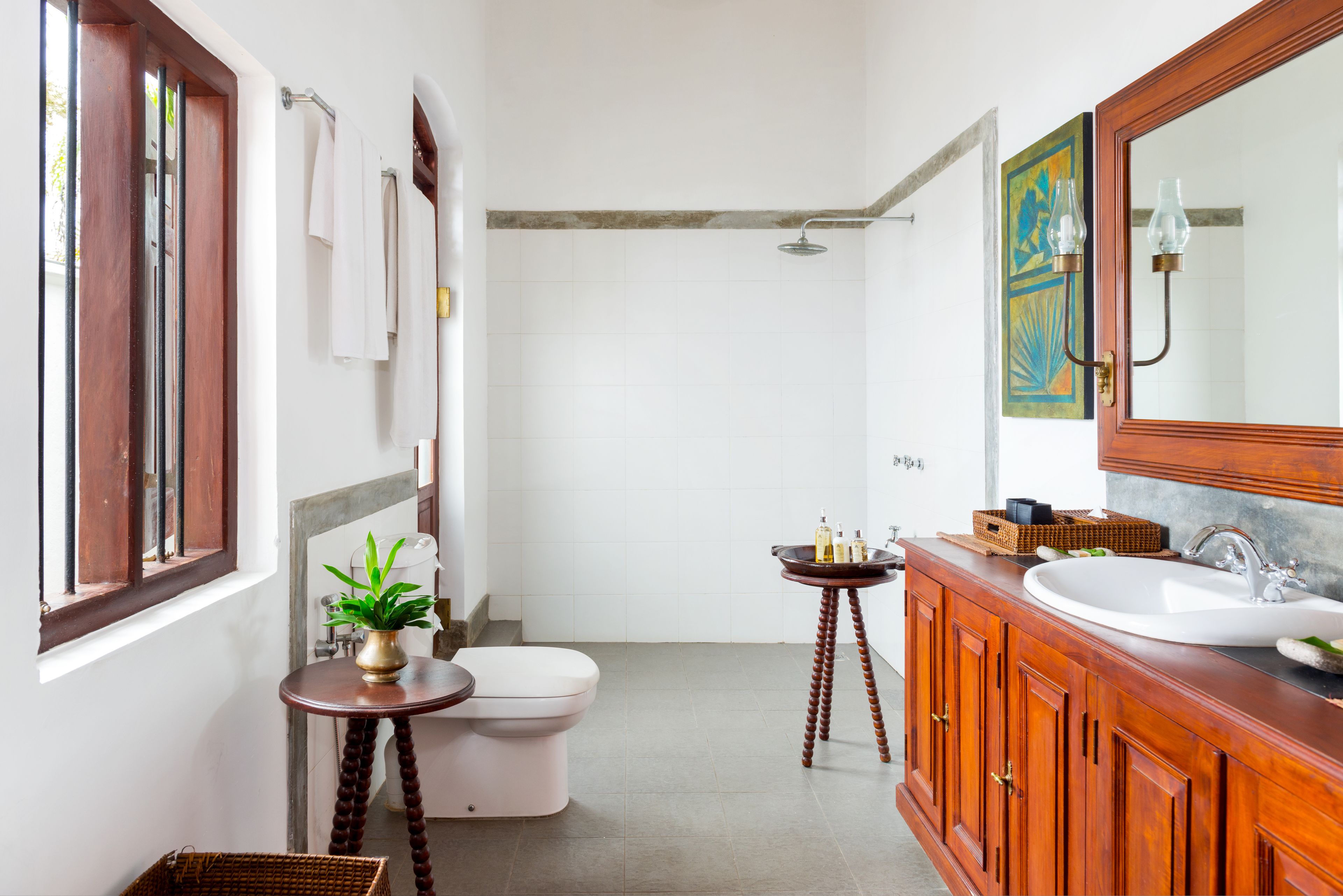 Un baño con inodoro, lavamanos y ducha. Hay una ventana, un espejo y una planta sobre una mesita.