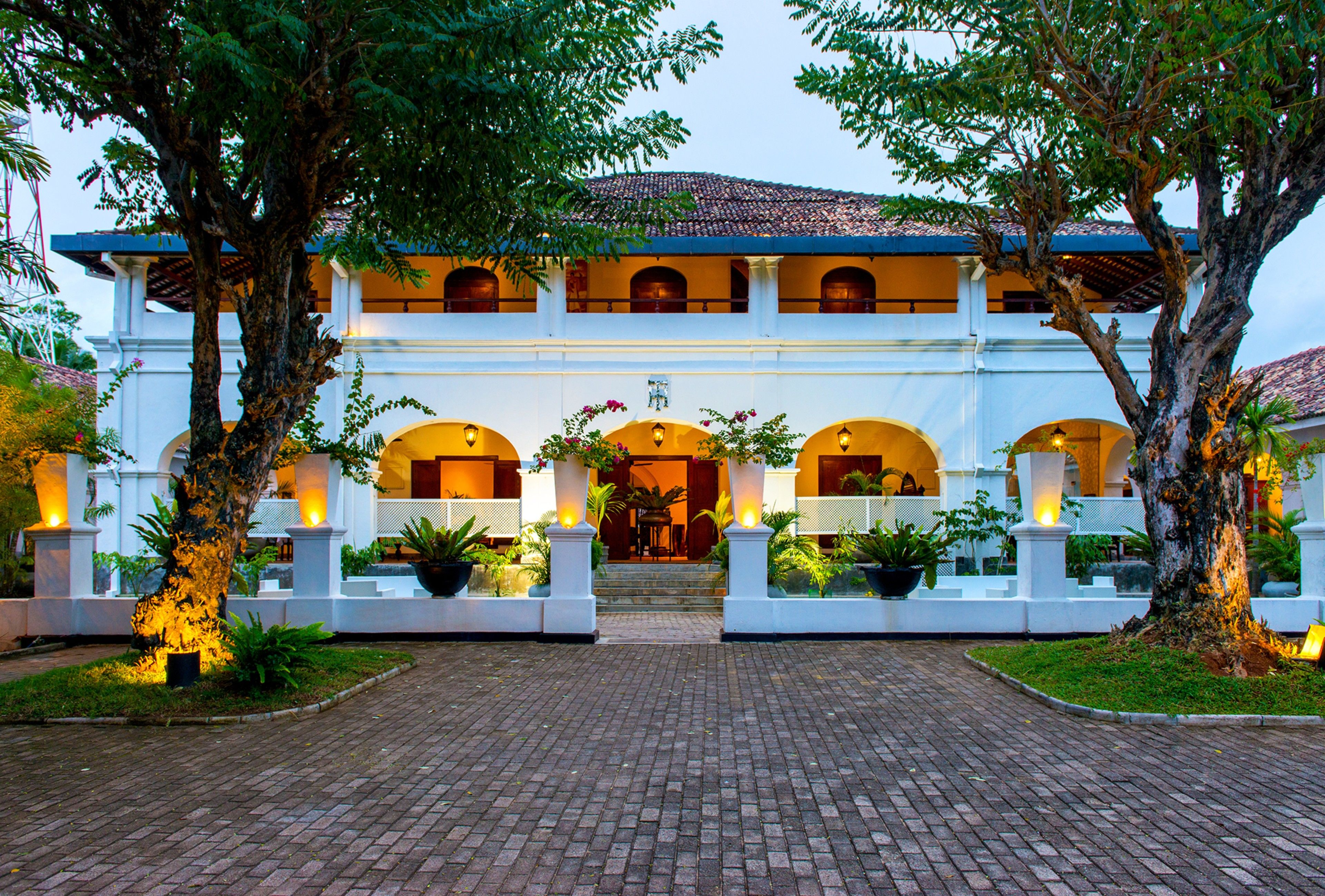 A large, white building with multiple arches and a tiled roof, surrounded by trees and potted plants.