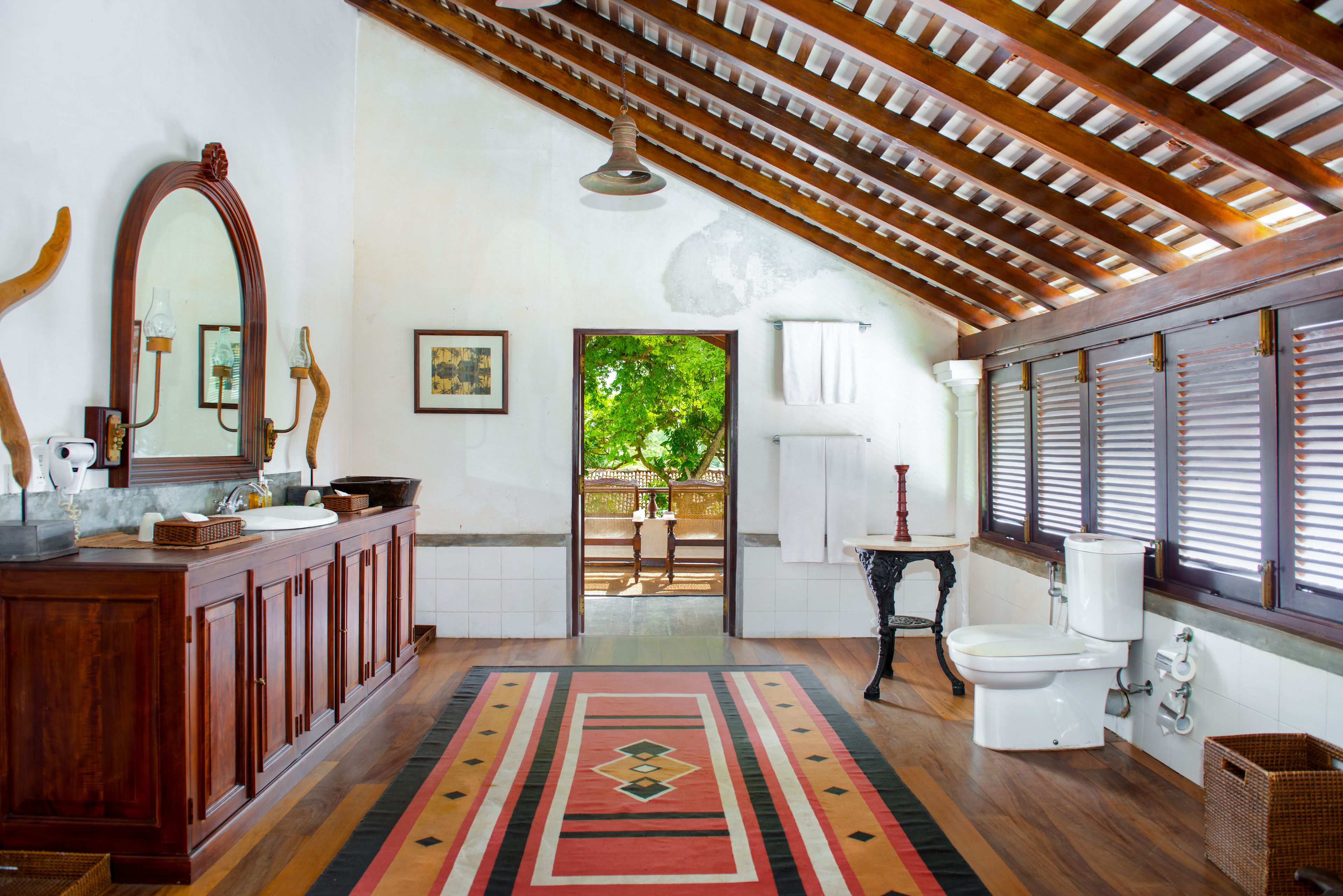A spacious bathroom with a wooden vanity, a large mirror, a toilet, and a window with shutters.