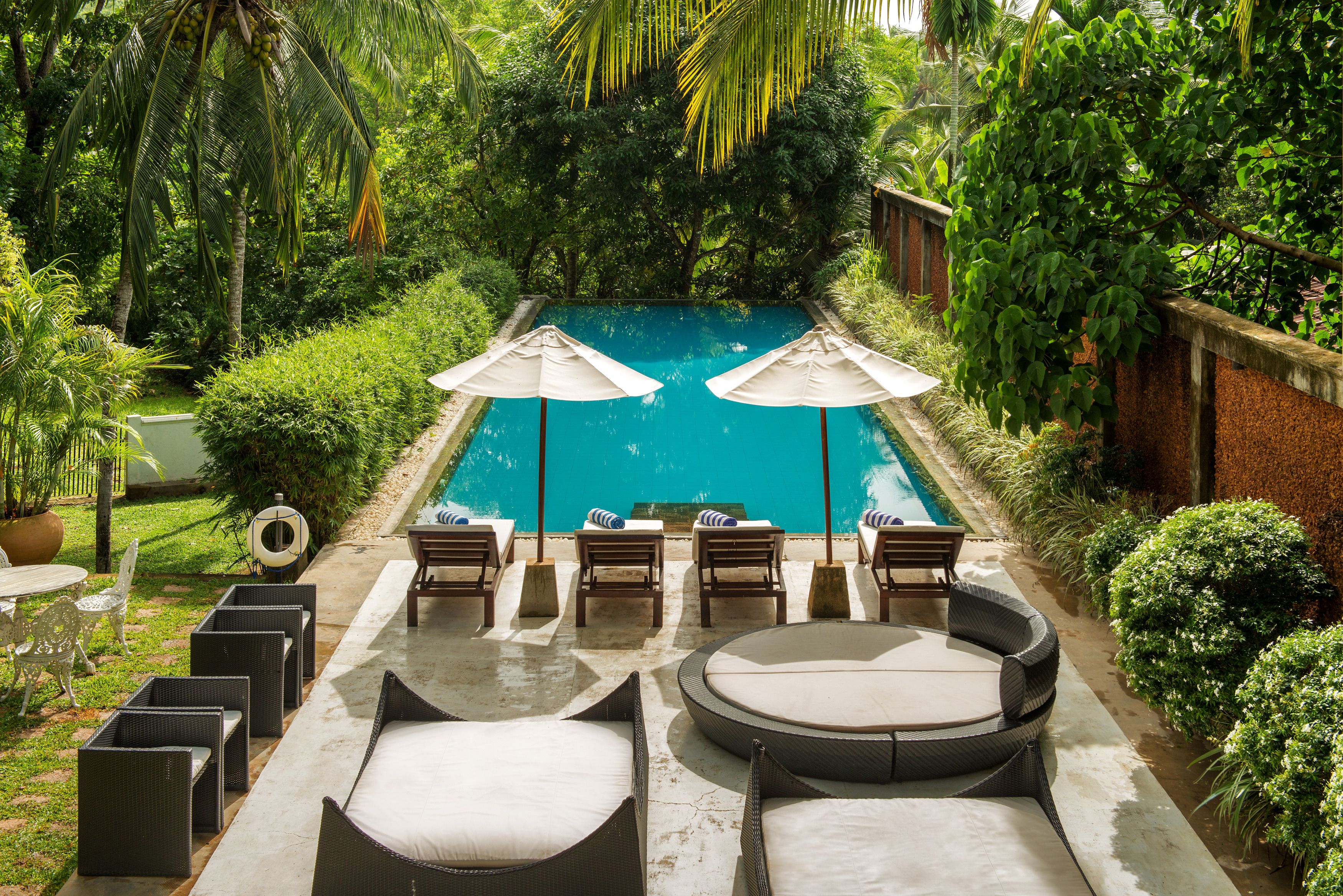 Zona de piscina con sillas de descanso y sombrillas, rodeada de exuberante vegetación.