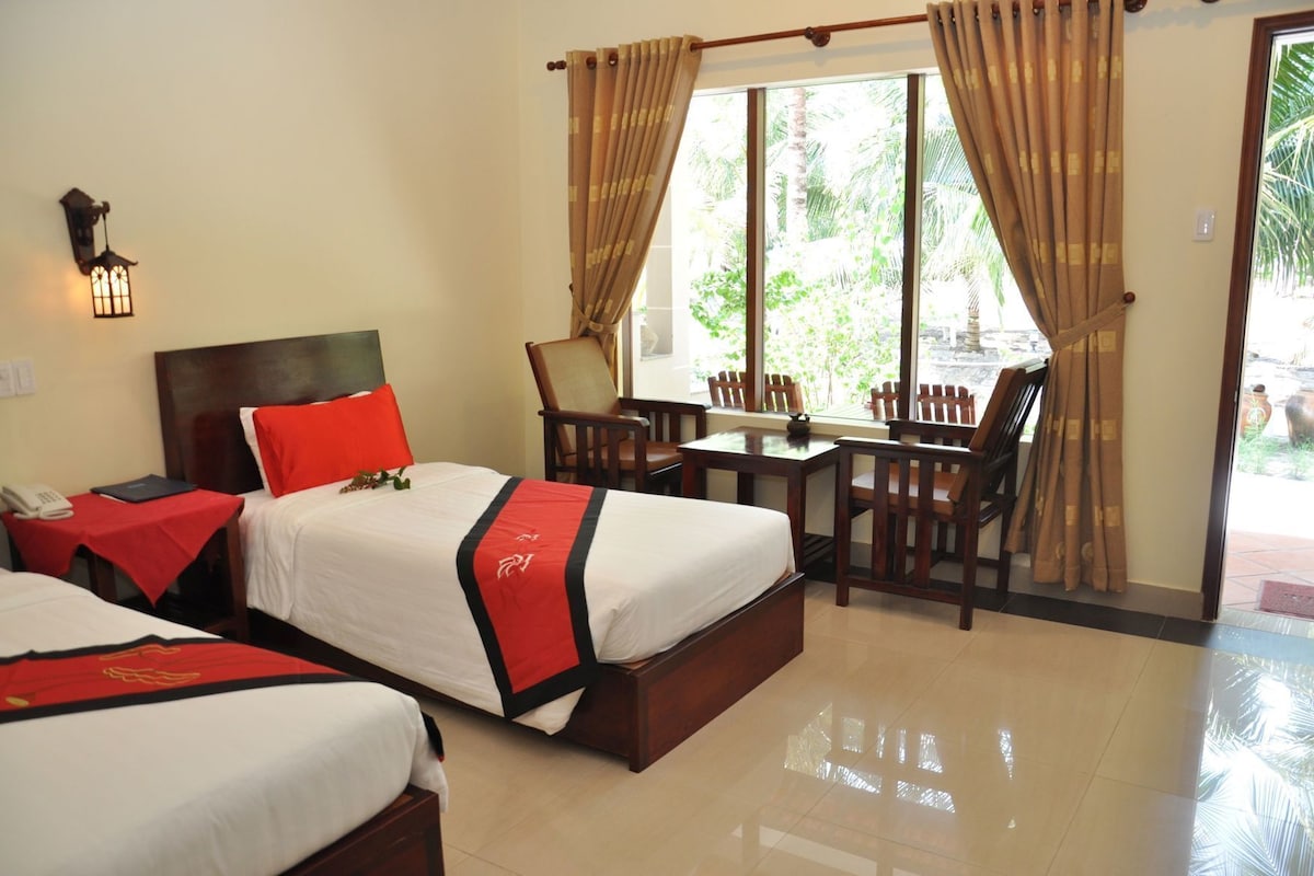 A hotel room with two beds, a table, chairs, a lamp, and a view of greenery outside.