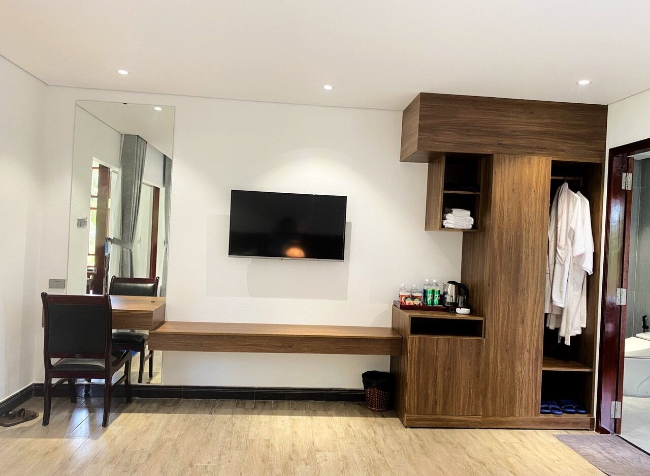 A modern hotel room with a wooden bench, a flat-screen TV mounted on the wall, a wooden wardrobe, and a mirror.