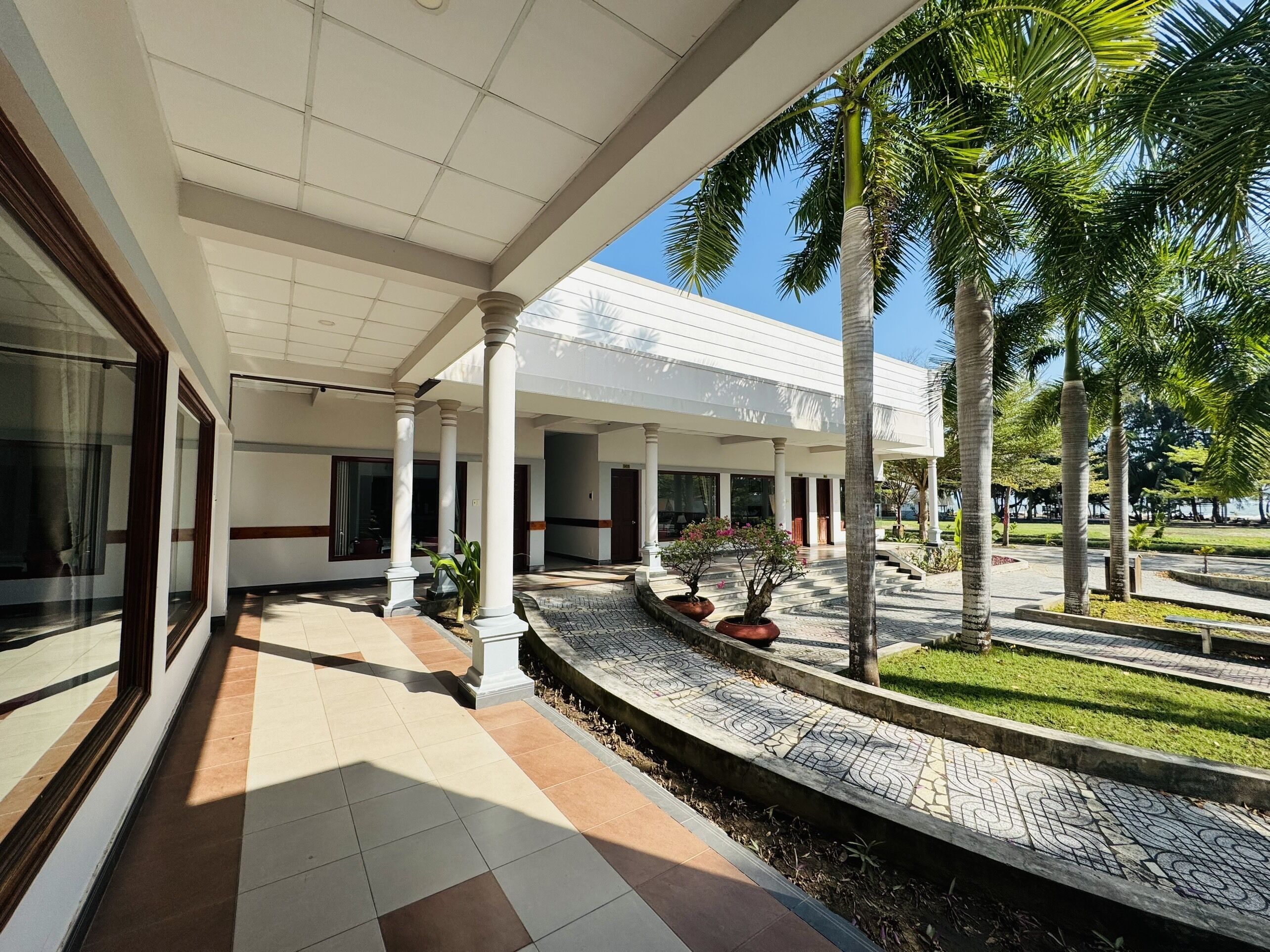 A covered outdoor area with a tiled floor, palm trees, and a building with large windows and columns.