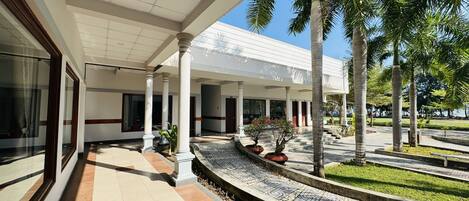 Un espacio al aire libre cubierto con suelo de baldosas, palmeras y un edificio con grandes ventanales y columnas.